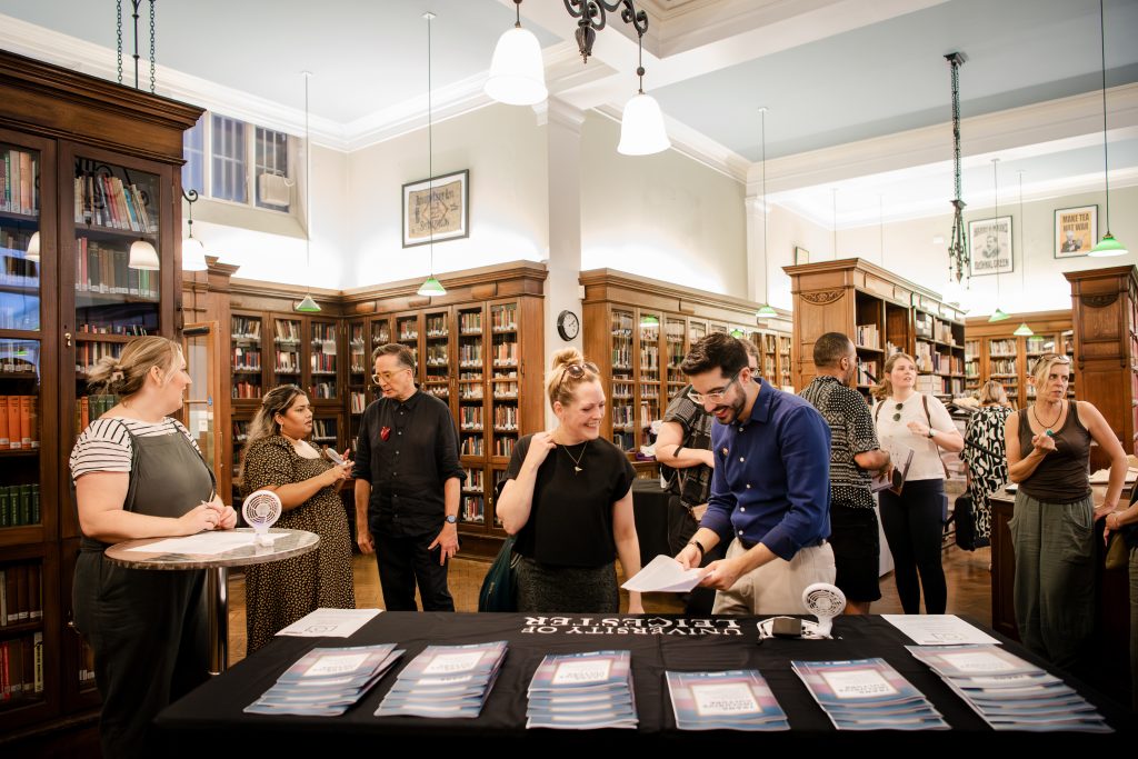 Room with many bookshelves, full of people chatting. Two people are speaking in front of a table with stacks of booklets.